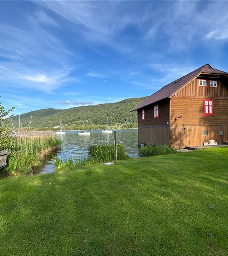 Ein idyllisches Holzhaus am Wasser, umgeben von grünem Gras und sanften Hügeln. Der Himmel ist klar und blau mit leichten Wolken.