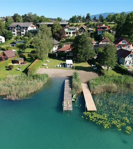 Ein ruhiger See mit einem kleinen Steg und üppigem Pflanzenbewuchs. Im Hintergrund sind gemütliche Häuser und ein klarer blauer Himmel zu sehen.