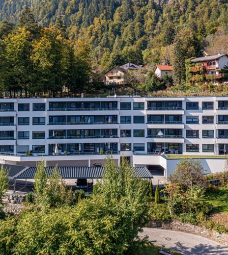 Un edificio moderno con più piani circondato da alberi e un giardino ben curato. Il cielo è sereno e il paesaggio è verde e accogliente.