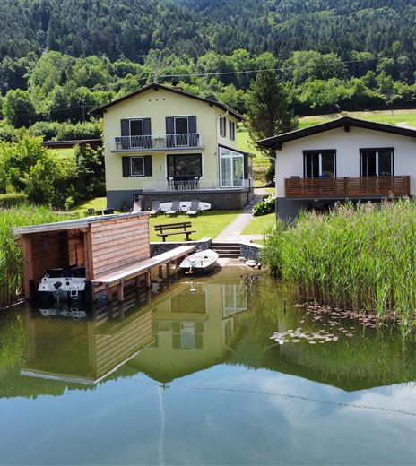 Eine ruhige Landschaft mit mehreren Häusern am Wasser. Das Ufer ist von hohen Pflanzen umgeben und spiegelt die Gebäude im klaren Wasser wider.