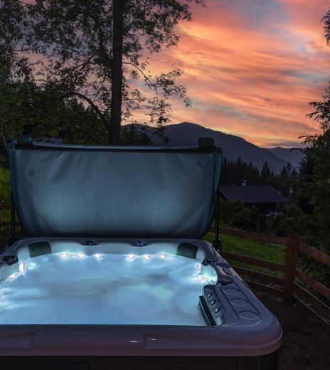 An outdoor hot tub surrounded by trees and a picturesque landscape. The sunset shines in warm colors over the horizon.