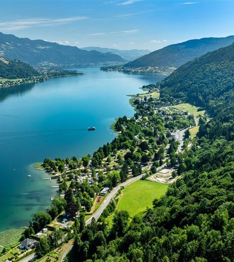 A beautiful lake surrounded by green mountains and forests. In the foreground are some houses and a paved path.