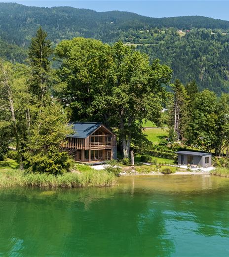 Ein wunderschönes Holzhaus am Ufer eines klaren, grünen Sees, umgeben von Bäumen. Im Hintergrund sind sanfte Hügel zu sehen.