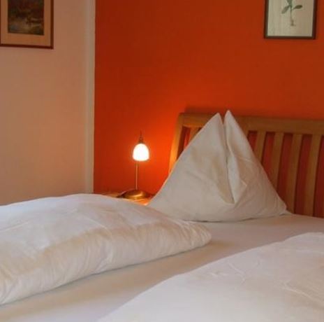 Ein gemütliches Schlafzimmer mit einem Holzbett und weißen Kissen. Die Wände sind in warmem Orange gestrichen, und es gibt sanftes Licht von einer Lampe.