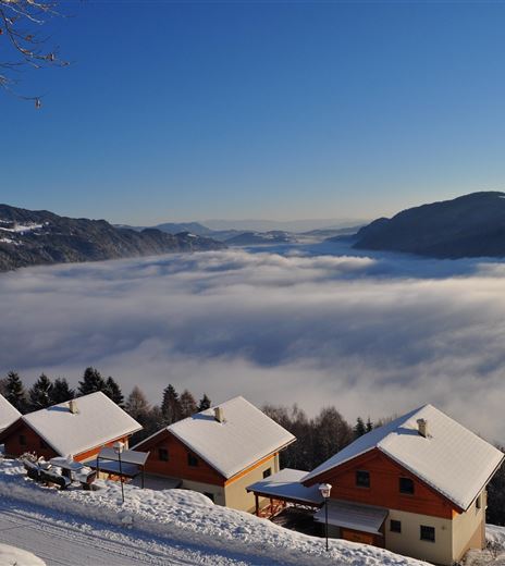 A snow-covered landscape with cozy huts. Below, a blanket of fog stretches between the mountains.