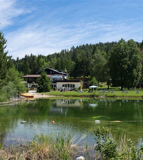Un tranquillo lago balneabile circondato da alberi e prati. Sullo sfondo si può vedere un edificio che invita a rilassarsi.