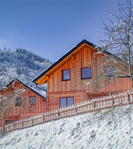 Zwei moderne Holzferienhäuser liegen malerisch in der verschneiten Landschaft. Im Hintergrund sind schneebedeckte Berge und ein klarer, blauer Himmel zu sehen.