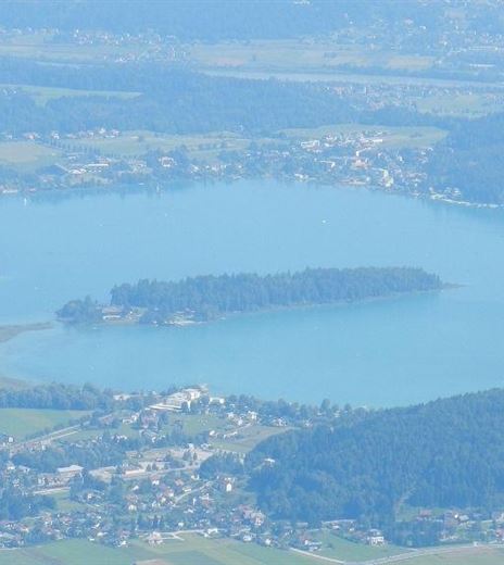 A beautiful view of a lake surrounded by green forests and small villages. The calm surface of the water reflects the landscape.