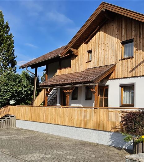Ein modernes Holzhaus mit einer hellen Fassade und einem geräumigen Balkon. Umgeben von grünen Bäumen und Blumenbeeten.