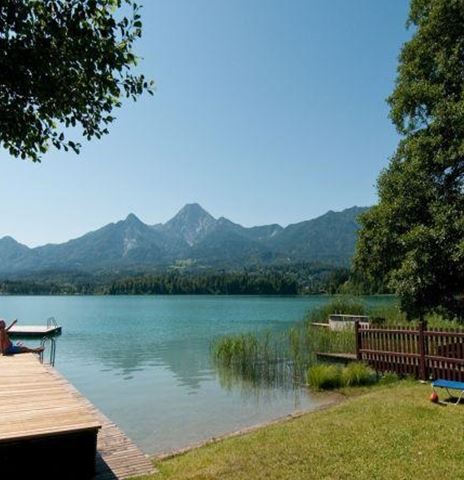 Ein ruhiger See mit Bergen im Hintergrund. Eine Holzterrasse am Wasser lädt zum Entspannen ein.