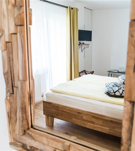 A cozy bedroom with a large bed and a wooden frame mirror. The windows are bright and decorated with yellow curtains.