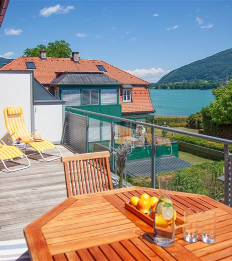 Una terrazza soleggiata con mobili in legno e vista su un lago limpido. Sullo sfondo si possono vedere montagne verdi e case incantevoli.