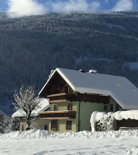 Ein schneebedecktes Landschaftsbild mit Häusern im Vordergrund. Im Hintergrund sind verschneite Berge und ein klarer blauer Himmel zu sehen.