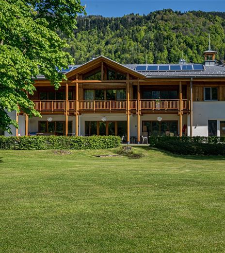 Un edificio moderno con rivestimento in legno e un ampio balcone. Circondato da erba verde e alberi in un paesaggio montano.