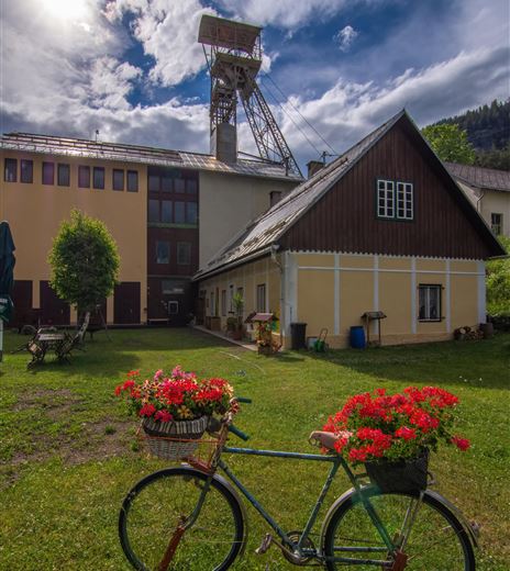 Ein rustikales Fahrrad mit blühenden roten Geranien steht auf einer Wiese. Im Hintergrund sind alte Gebäude und ein Förderturm zu sehen.