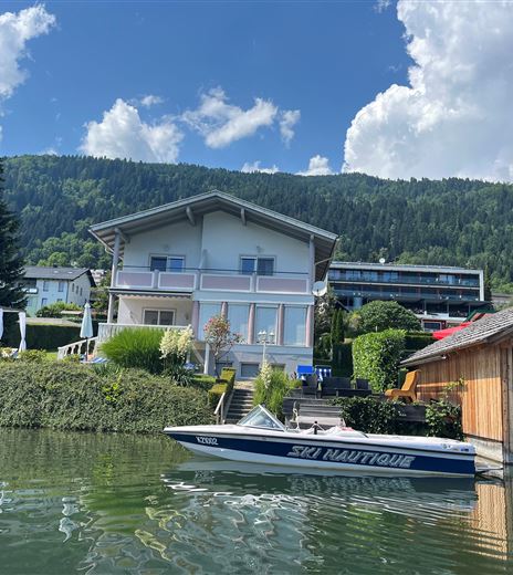Una bella casa sull'acqua con una barca e un ambiente verde. Le montagne sullo sfondo e le nuvole nel cielo conferiscono alla scena un'atmosfera rilassata.