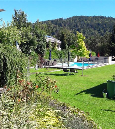 Ein schöner Garten mit gepflegtem Rasen und bunten Pflanzen. Im Hintergrund befindet sich ein Swimmingpool, umgeben von Bäumen und Bergen.