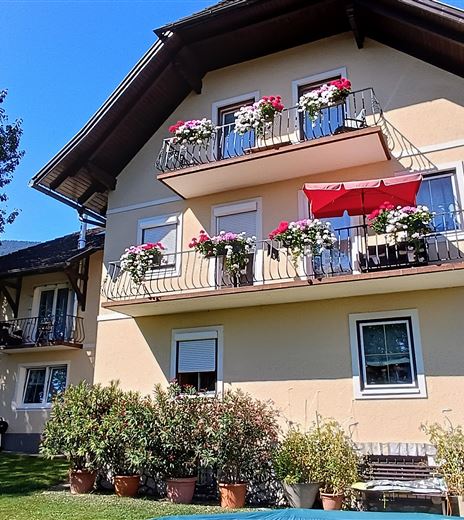 Ein hübsches Gebäude mit einem Balkon, der mit Blumen dekoriert ist. Im Vordergrund gibt es Pflanzen und Bäume, die eine einladende Atmosphäre schaffen.