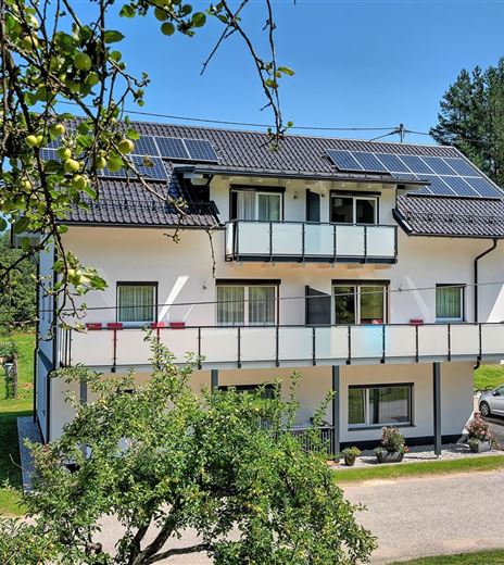 Ein modernes Haus mit Solarzellen auf dem Dach und einem Balkon. Umgeben von Grünflächen und Bäumen.