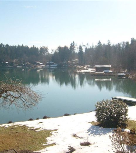 Ein ruhiger See mit klarem Wasser und Schnee am Ufer. Im Hintergrund sind Bäume und einige Gebäude sichtbar.