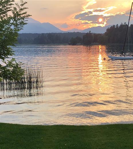 Ein ruhiger See mit funkelndem Wasser und einer malerischen Abenddämmerung. Im Hintergrund sind leicht bewaldete Hügel und ein Segelboot zu sehen.