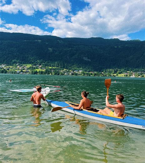 Three people in the water on kayaks, surrounded by mountains and a clear sky. It's a sunny day and the atmosphere is relaxed.