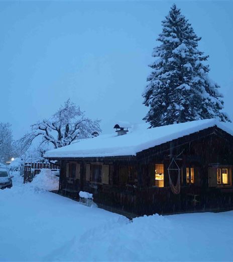 A cozy wooden cabin in winter, surrounded by fresh snow. The windows glow warmly in the blue twilight.