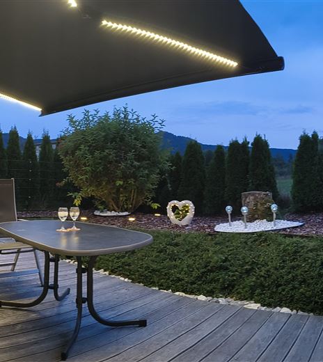 Ein gemütlicher Außenbereich mit einem Tisch und Stühlen unter einem Sonnenschutz. Der Garten ist grün mit einigen Pflanzen und einer malerischen Aussicht.