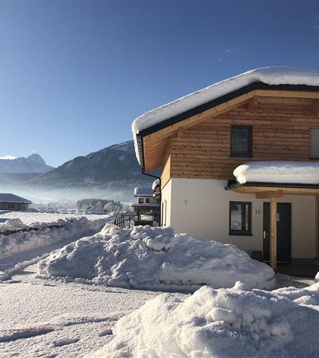 A modern wooden house in the snow with mountains in the background. The landscape is tranquil and wintry.