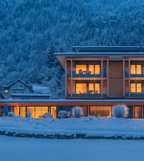 Ein modernes Gebäude in einer schneebedeckten Landschaft, beleuchtet von warmem Licht. Die Umgebung ist ruhig und winterlich, umgeben von Bäumen.