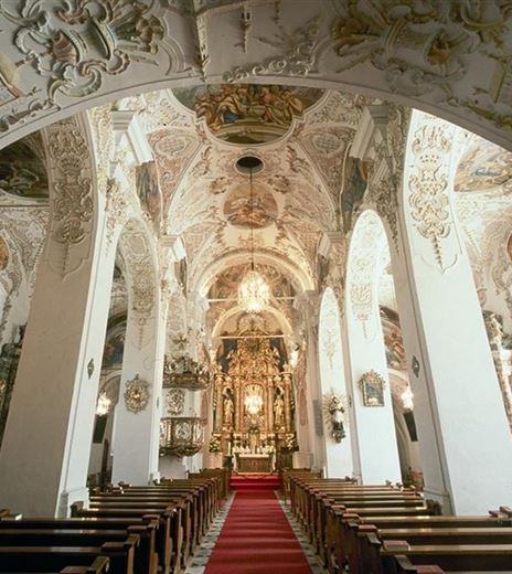 An impressive church with elaborate ceiling paintings and a red carpet in the interior. The benches are neatly lined up and lead to the altar area.