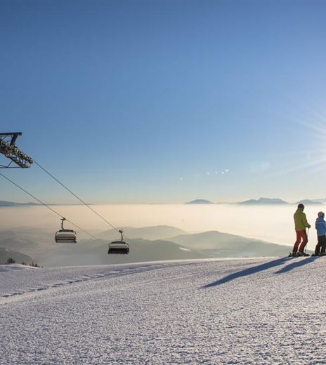 A snowy mountain landscape with a cable car and two skiers. The sky is clear and the sun is shining brightly.