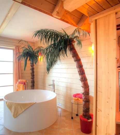 Ein modernes Badezimmer mit einer freistehenden Badewanne und Palmen. Im Hintergrund befindet sich eine Sauna und große Fenster, die viel Licht hereinlassen.