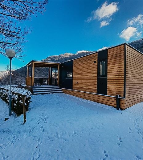 Una moderna casa in legno nella neve con un cielo blu chiaro. Circondata da montagne coperte di neve e alberi.