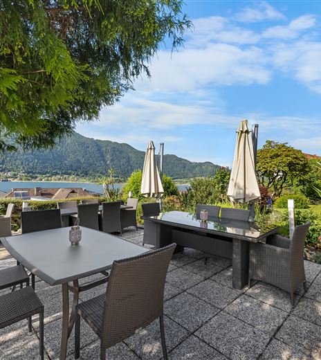 Eine gemütliche Terrasse mit einem Tisch und Stühlen. Im Hintergrund sind der See und die Berge sichtbar, umgeben von Pflanzen.