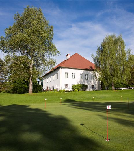 Ein gepflegter Golfplatz mit einem malerischen Schloss aus dem 17. Jahrhundert im Hintergrund. Hohe Bäume umgeben die grüne Landschaft unter einem klaren Himmel.