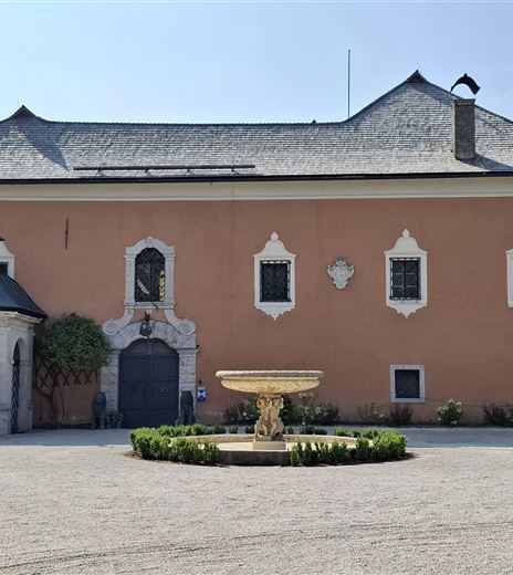 Un edificio storico con una facciata rossa e un bel giardino. In primo piano c'è una fontana, circondata da aiuole fiorite.