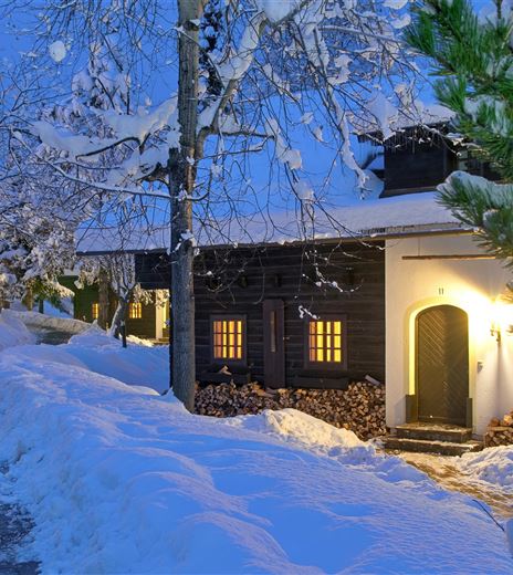 Ein winterliches Dorf mit schneebedeckten Bäumen und einem gemütlichen Holzhaus. Sanftes Licht strahlt aus den Fenstern und beleuchtet den verschneiten Weg.