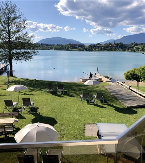 Un paesaggio idilliaco al lago con un prato verde, lettini e ombrelloni. Sullo sfondo si vedono le montagne e un cielo blu chiaro.