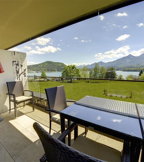 Una bellissima terrazza con un tavolo e sedie si affaccia su un paesaggio pittoresco. Sullo sfondo si vedono montagne e un lago tranquillo, sotto un cielo blu con alcune nuvole.