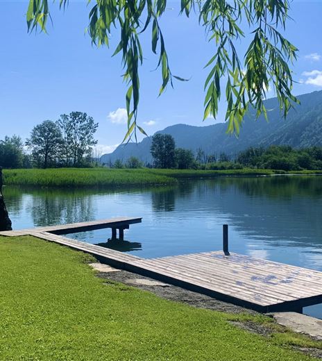 Ein ruhiger See umgeben von grünen Wiesen und Bergen. Ein Holzsteg führt ins Wasser.