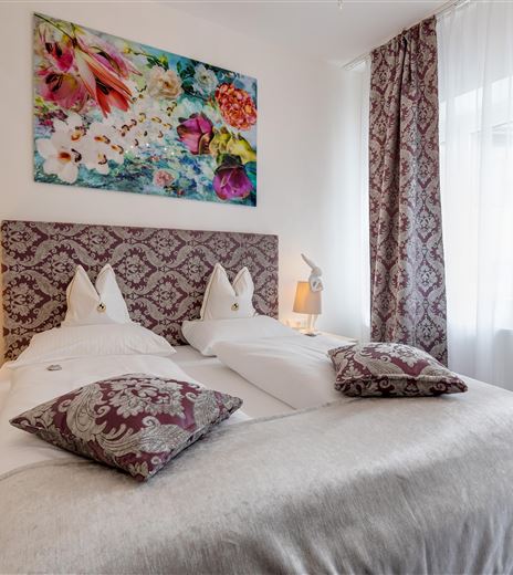 Ein stilvolles Schlafzimmer mit einem großen Bett und dekorativen Kissen. Die Wände sind mit ansprechenden Vorhängen und einem bunten Wandbild geschmückt.