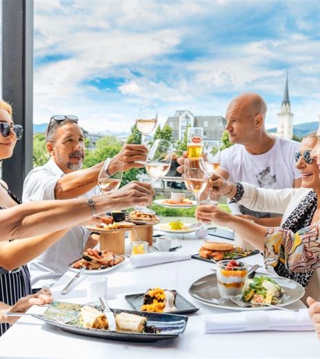 Un gruppo di persone alza i bicchieri e festeggia durante un pasto all'aperto. Sullo sfondo ci sono volti sorridenti e una vista accattivante.