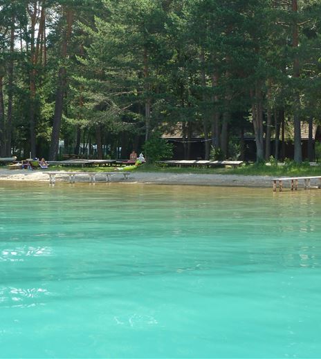 Un lago cristallino di colore turchese con una spiaggia di sabbia. Sullo sfondo si vedono alberi e sdraio.