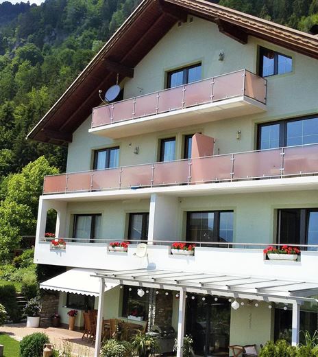 Una casa moderna con diversi balconi e un bel giardino. L'ambiente è circondato da montagne e tanto verde.
