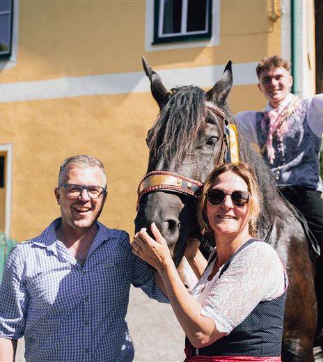 A group of three people poses with a black horse in front of a yellow house. Two people stand by the horse, while one person sits on it and smiles.