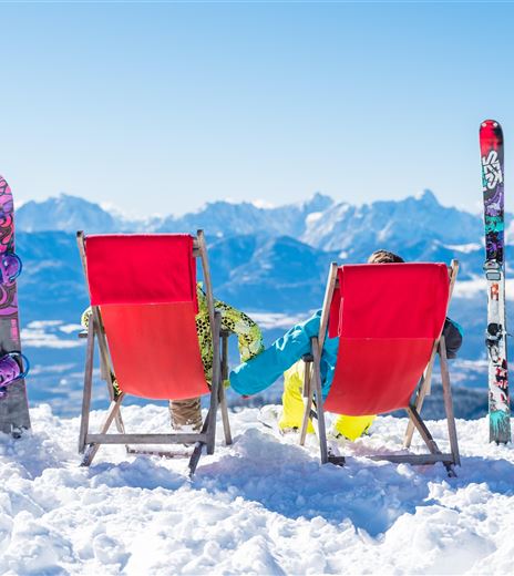 Due sdraio rosse sono nella neve con vista sulle montagne. Accanto a loro si appoggiano tavole da neve e da sci, mentre il sole splende.