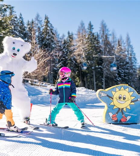 due bambini in abbigliamento da sci stanno sulla pista e sorridono, mentre un grande orso polare amichevole li accompagna. Sullo sfondo si vedono un cielo azzurro e alberi innevati.