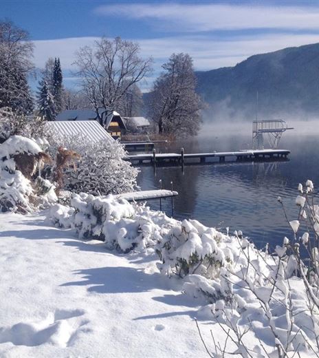 Paesaggio innevato al lago con un pontile in legno e un'atmosfera tranquilla. Da un lato si possono vedere alberi coperti di neve e una piccola baita.