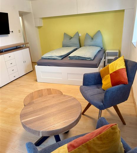 Ein gemütliches Zimmer mit einem Doppelbett und blauen Kissen. Es gibt einen runden Holztisch und einen Sessel mit bunten Kissen.